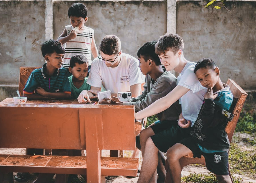 Racers showing a camera to Thai boys at a wooden table